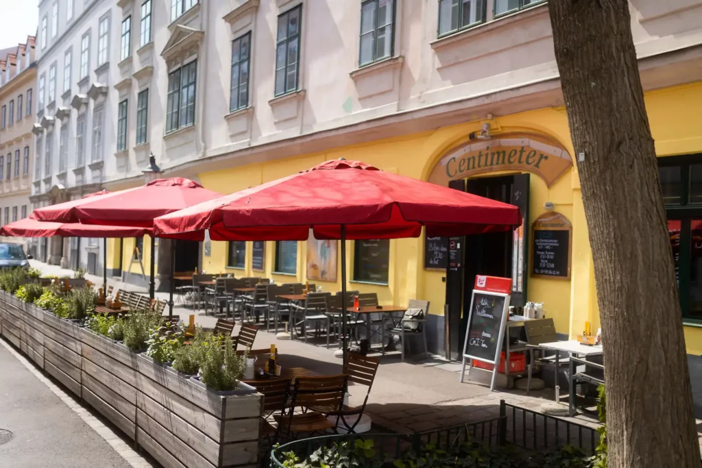 Außenansicht eines Restaurants Namens Centimeter mit roten Sonnenschirmen und Sitzplätzen im Freien im Gastgarten Wien, gemütlicher Gastgarten vor einem Lokal in einer Wiener Altbaustraße.