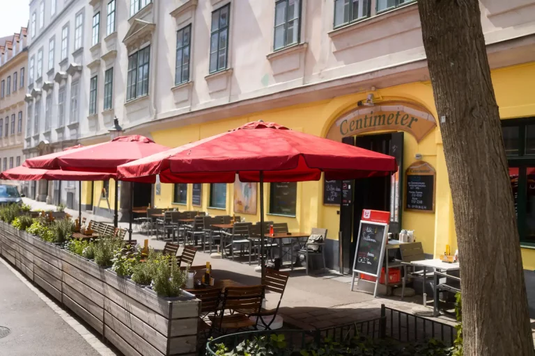 Außenansicht eines Restaurants Namens Centimeter mit roten Sonnenschirmen und Sitzplätzen im Freien im Gastgarten Wien, gemütlicher Gastgarten vor einem Lokal in einer Wiener Altbaustraße.