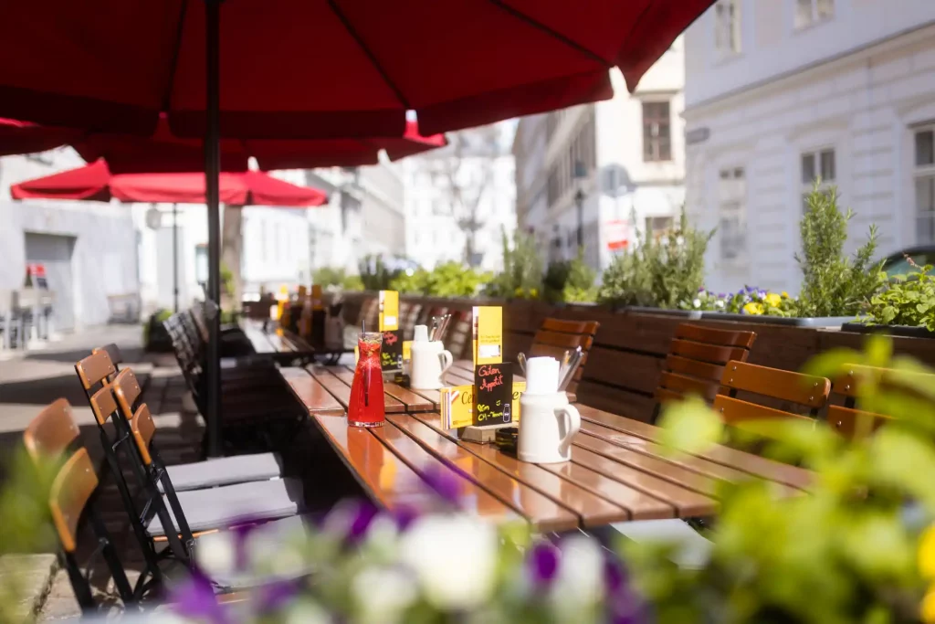 Außenansicht eines Restaurants Namens Centimeter mit roten Sonnenschirmen und Sitzplätzen im Freien im Gastgarten Wien, gemütlicher Gastgarten vor einem Lokal in einer Wiener Altbaustraße.